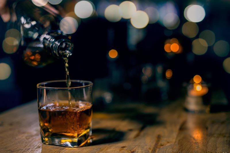 A person is pouring whiskey into a glass on a wooden table.