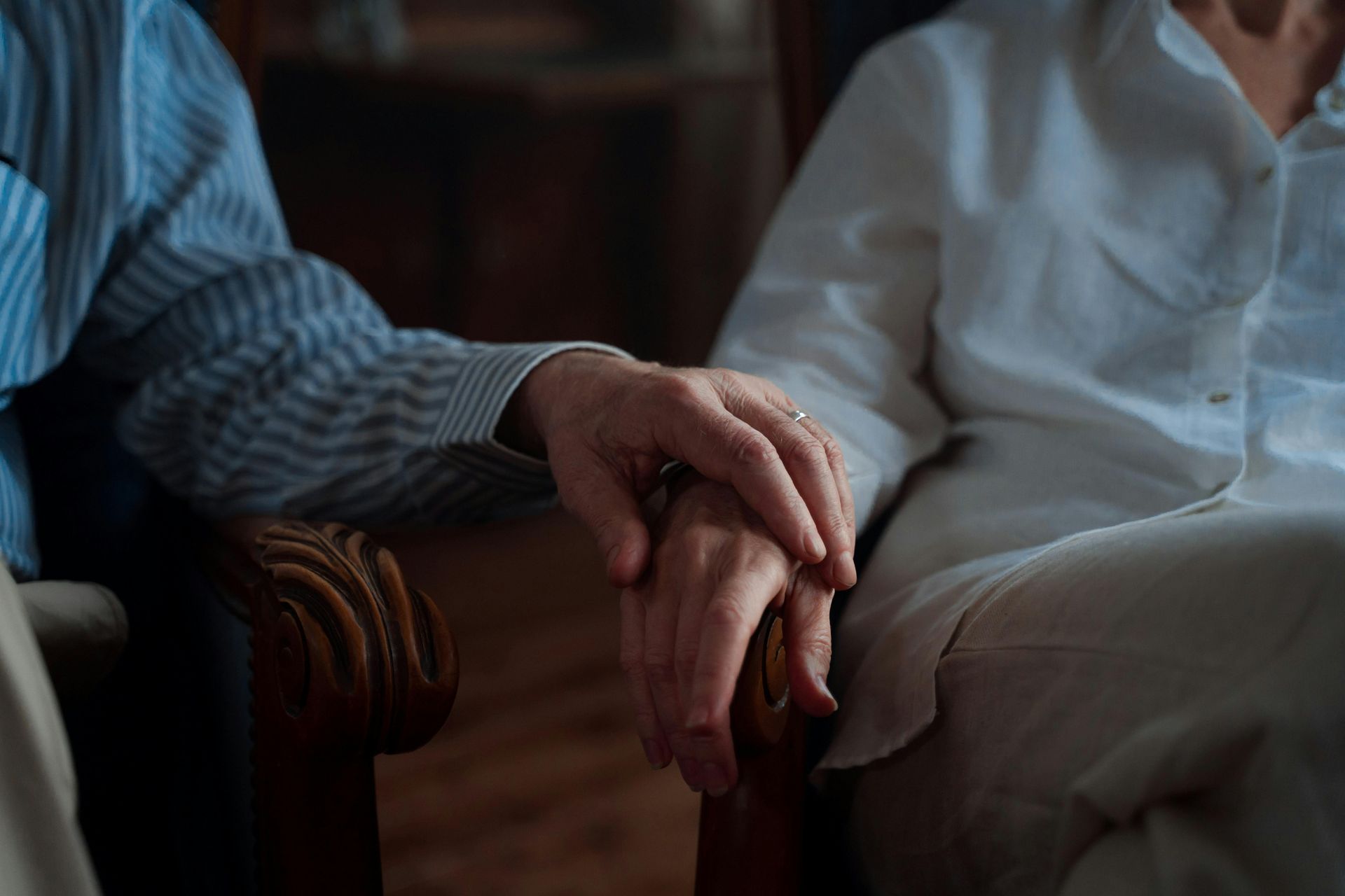 Two people hold hands, seated. One in striped shirt, the other in white, indoors.
