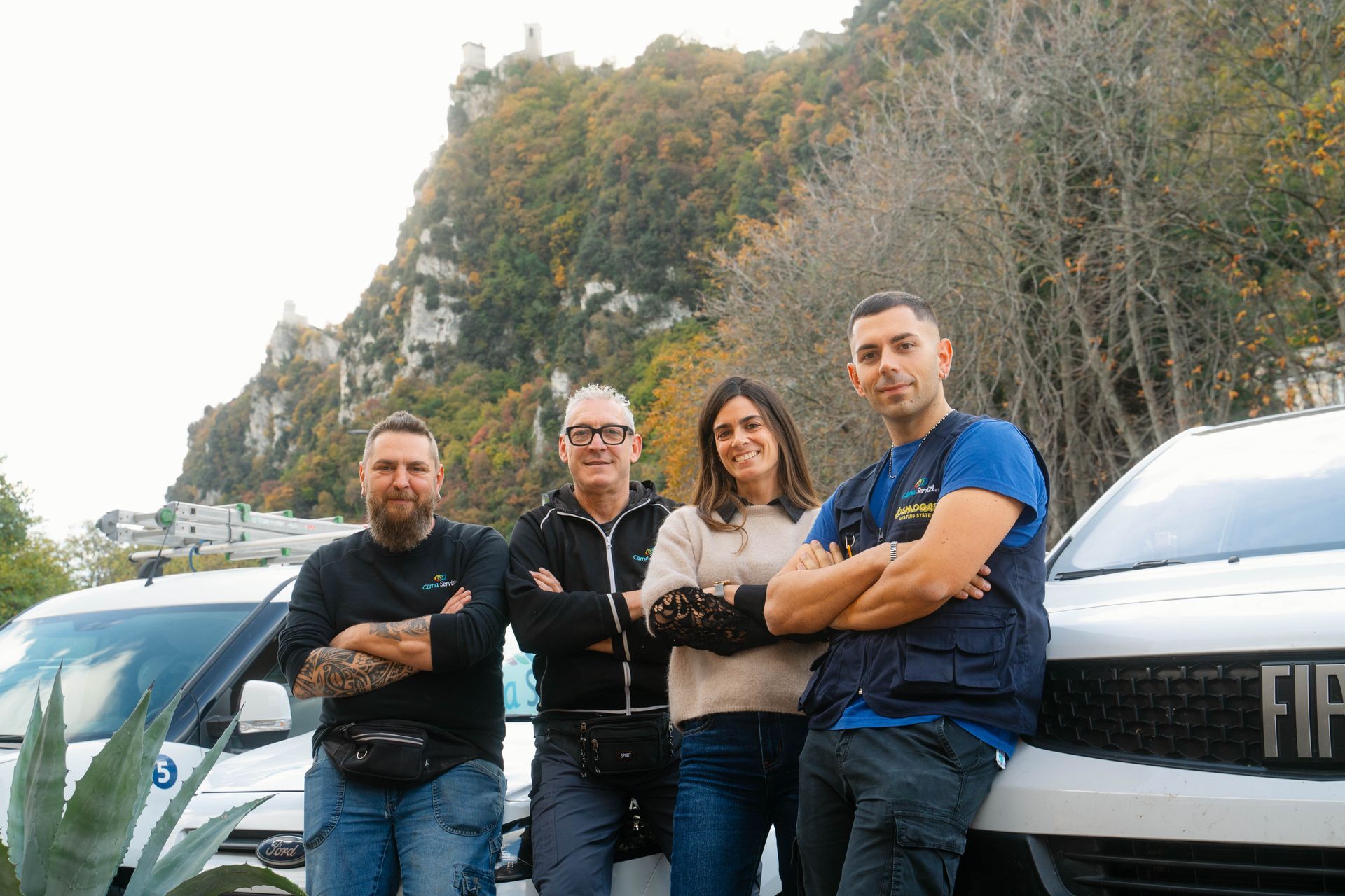 Quattro persone sono in piedi vicino ad auto bianche, con le montagne sullo sfondo. Una persona ha le braccia incrociate, l'altra sta toccando un'auto.