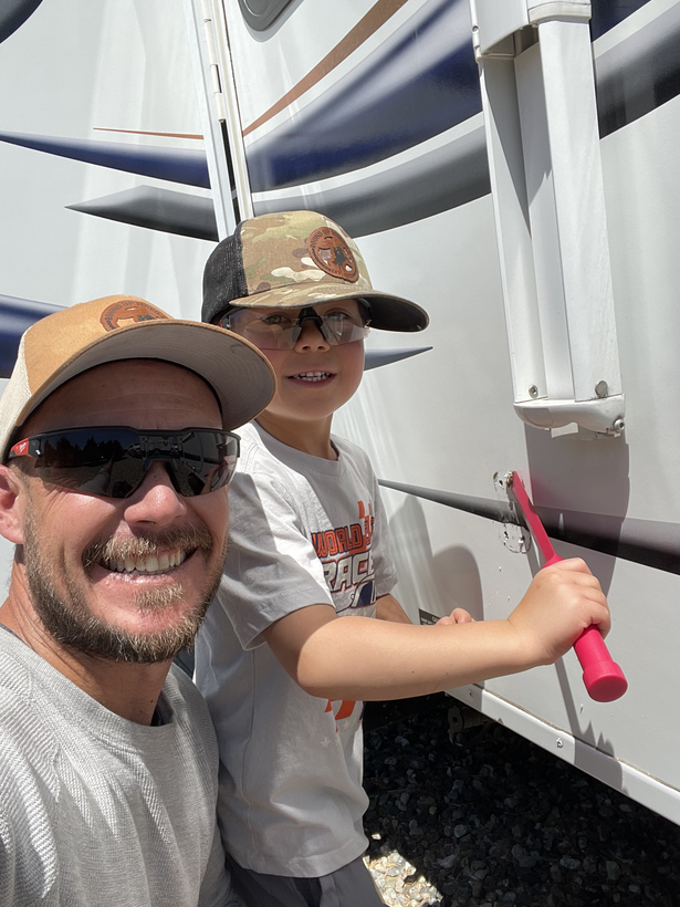 Man and child cleaning an RV with toothbrushes; sunny outdoor setting.