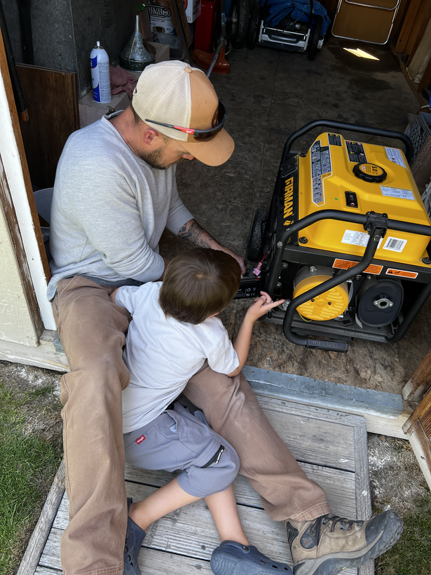 Man and child working on a yellow generator. The child sits in the man’s lap in a shed.