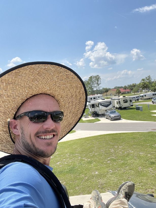 Man smiling, wearing sunglasses and straw hat, in a campground with RVs and blue sky.