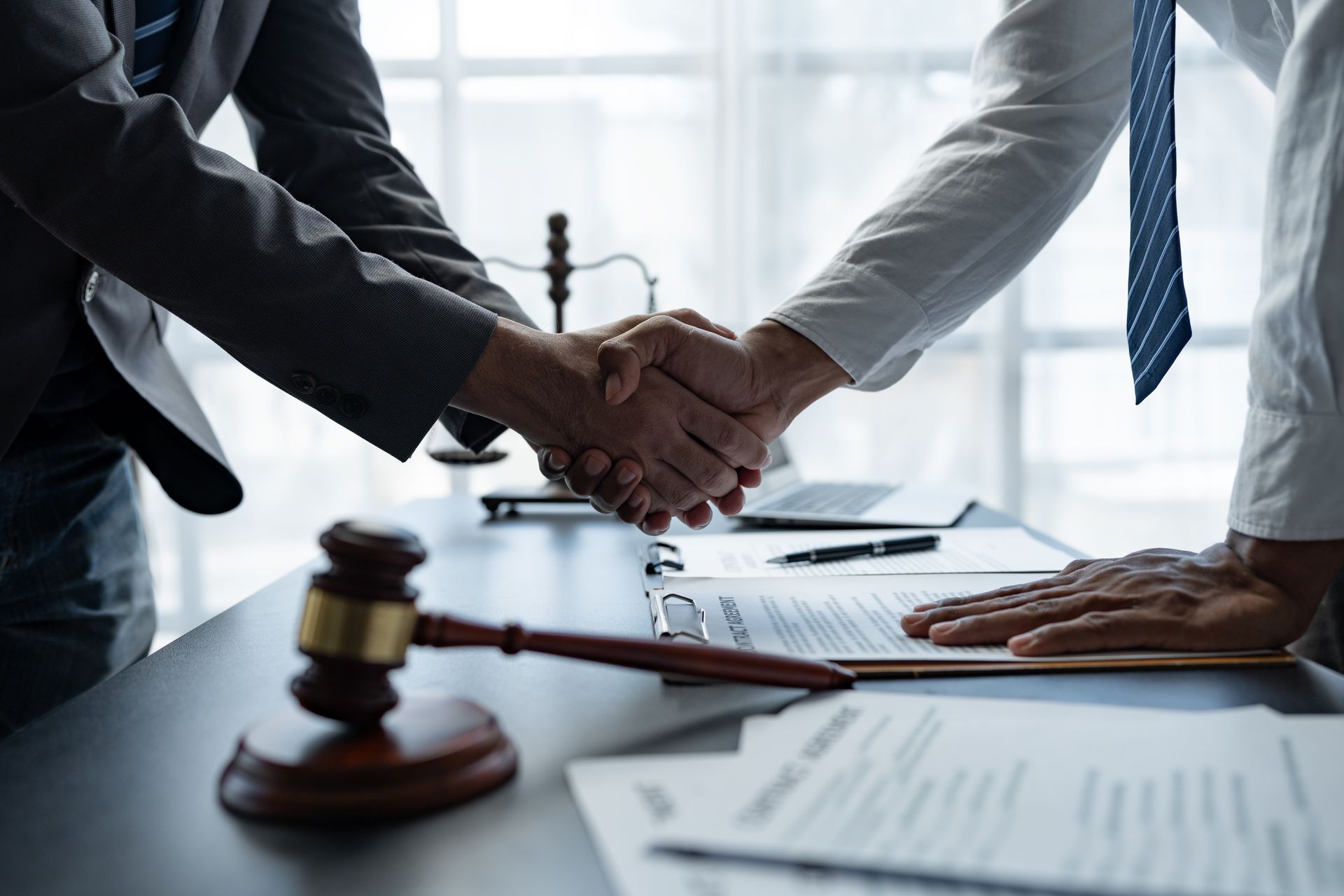 Business handshake over desk with gavel, documents, and laptop, suggesting a legal agreement