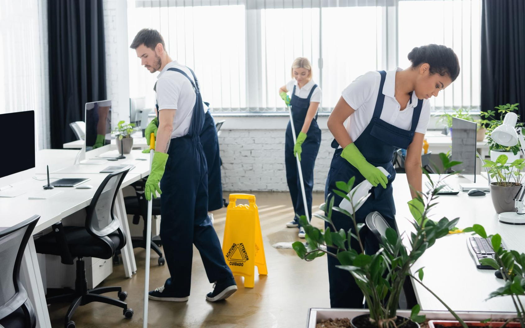People cleaning an office floor with mops. Yellow gloves and green pants are visible. Another person cleans glass in the background.