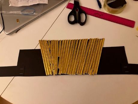 Craft table with black cardboard, yellow striped piece, scissors, and pink ruler on a light surface