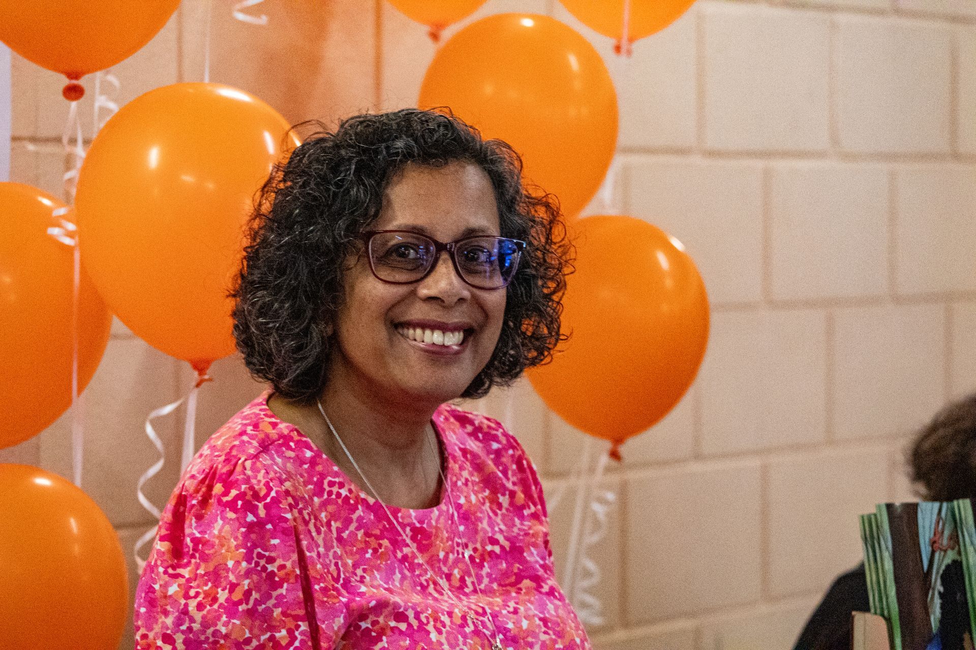 A woman wearing glasses and a pink shirt is smiling in front of orange balloons.