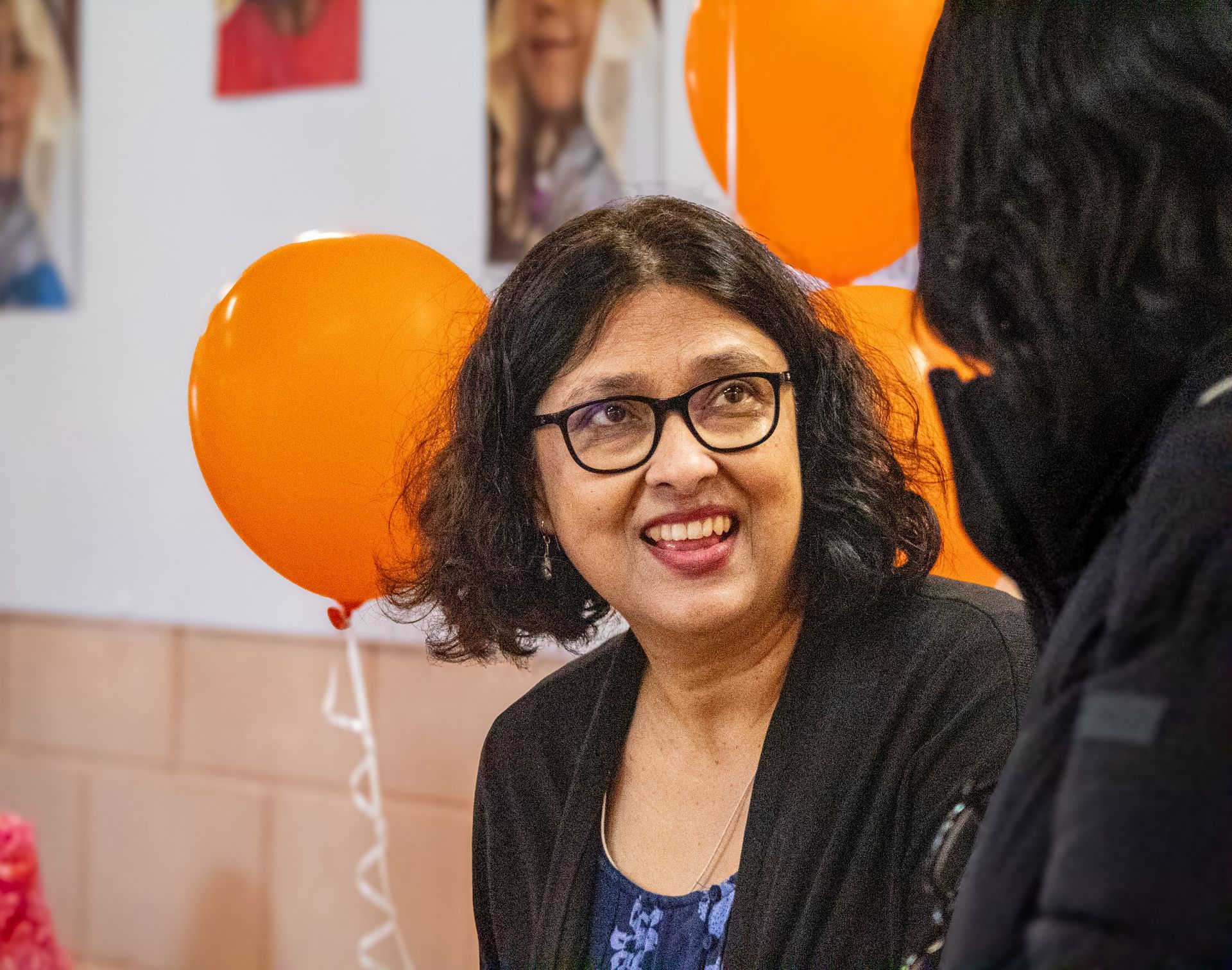 A woman wearing glasses is smiling while talking to another woman.