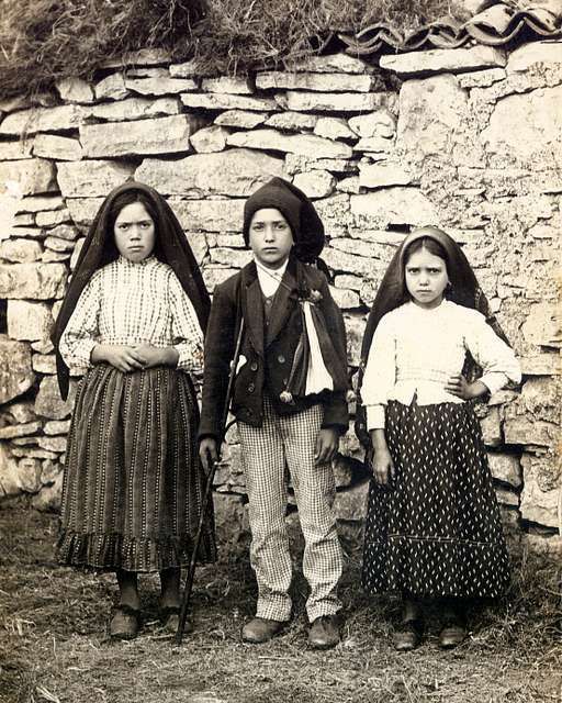Three children in traditional clothing standing in front of a stone wall.