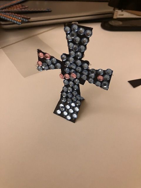 Small beaded black cross with pink accents standing on a beige tabletop, with notebooks in the background