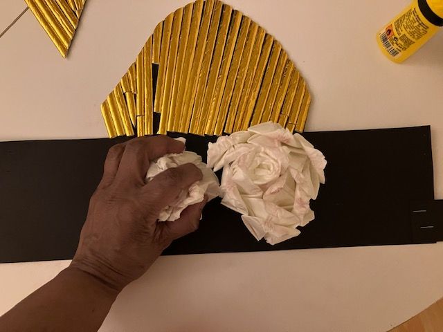 Hand arranging white tissue on a black strip with a gold fan-shaped craft piece on a table