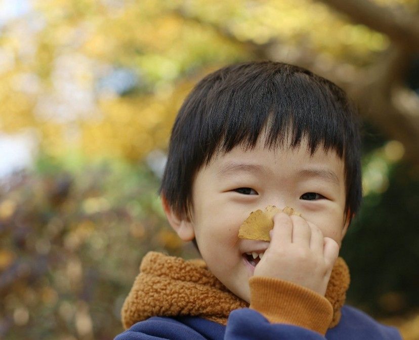 Smiling child holding a yellow leaf up to their nose outdoors, wearing a brown scarf and blue jacket.