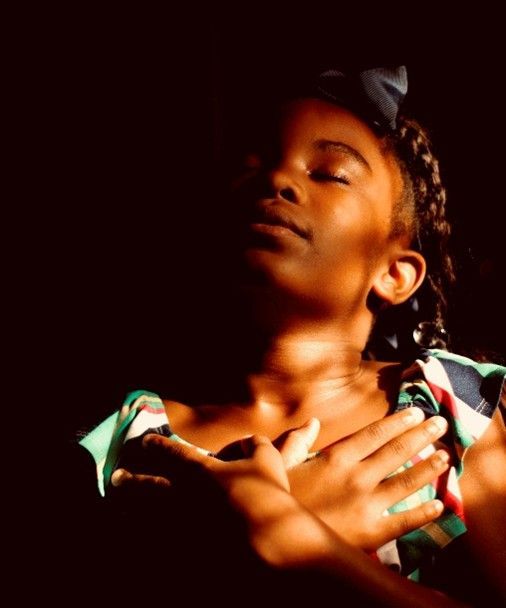 Young Black girl with eyes closed, hands on chest, bathed in sunlight.