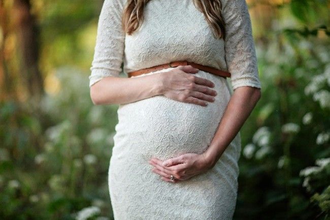 Pregnant woman in a white lace dress, hands on her belly, standing outdoors with green foliage.