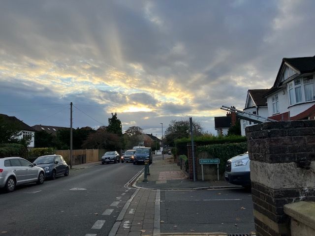 Street scene with parked cars, sidewalk, houses, and a cloudy sky with sun rays.