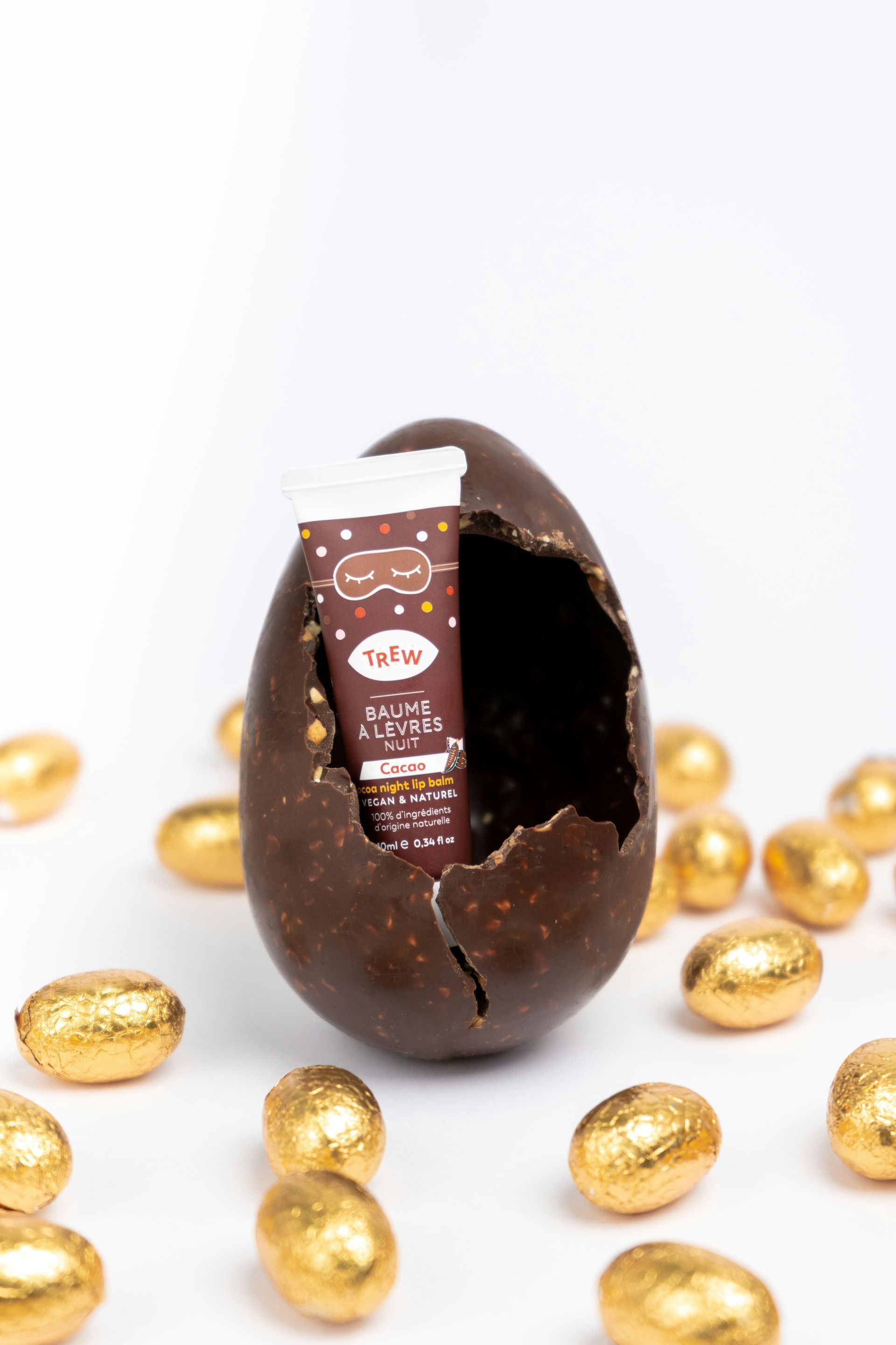 Chocolate Easter egg with skincare product, surrounded by golden eggs on white.
