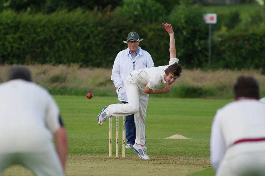 Cricket bowler in white uniform bowls, red ball in air, umpire watches, two batsmen in foreground.