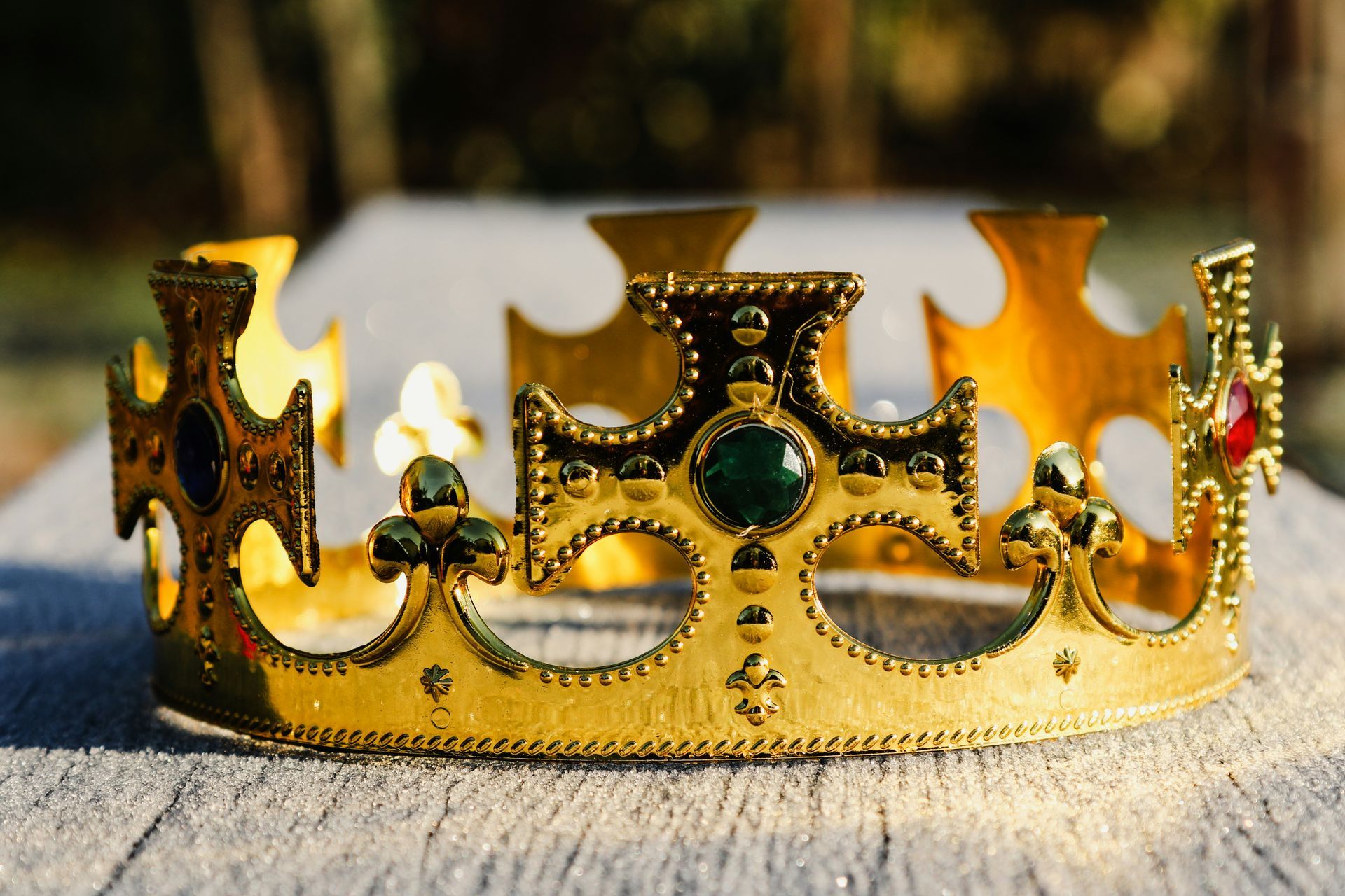 Gold crown with green and red jewels on a wooden surface outdoors, blurred background.