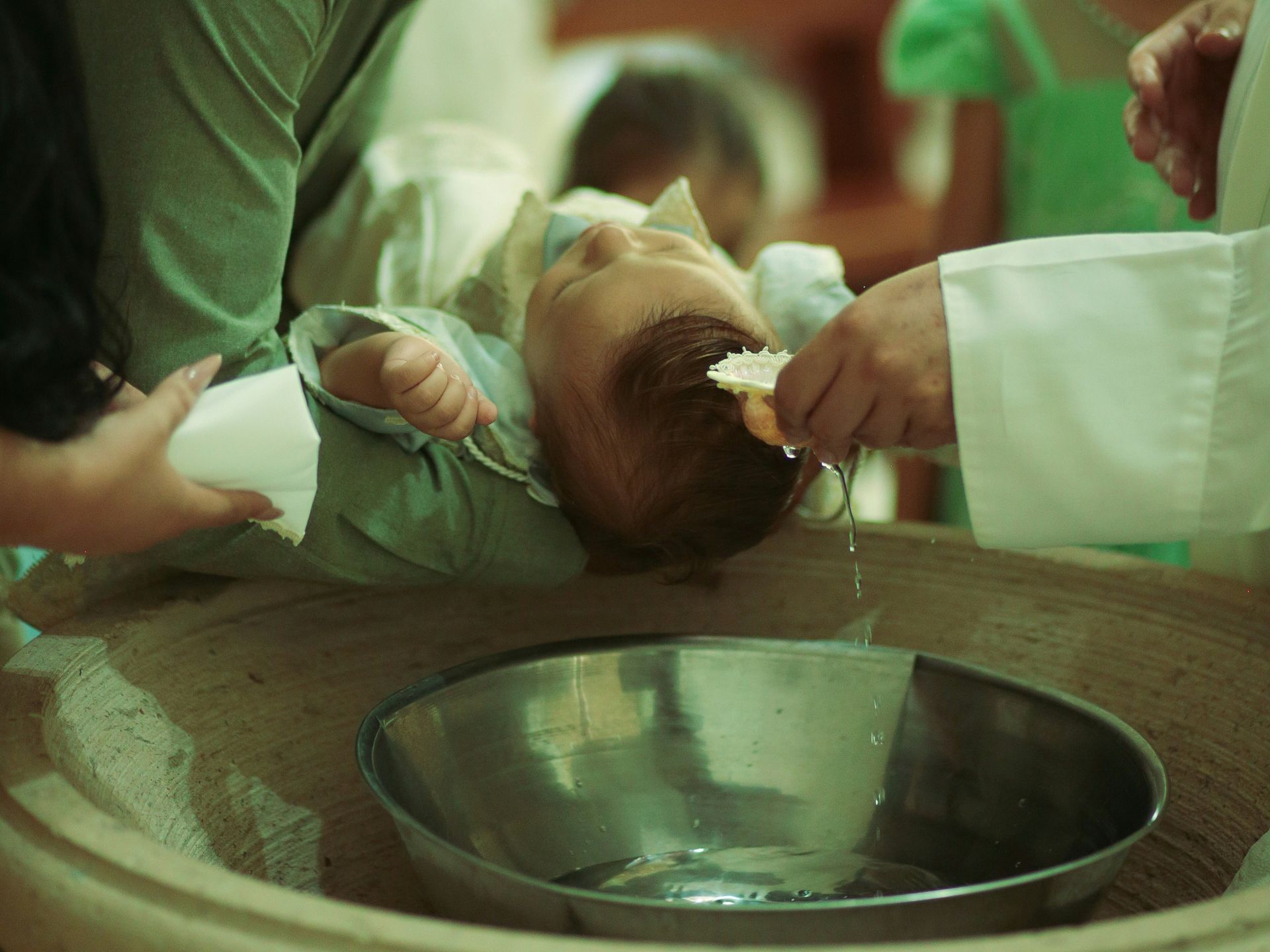 An infant receives a baptism as water is poured over their head from a small shell into a metal bowl.