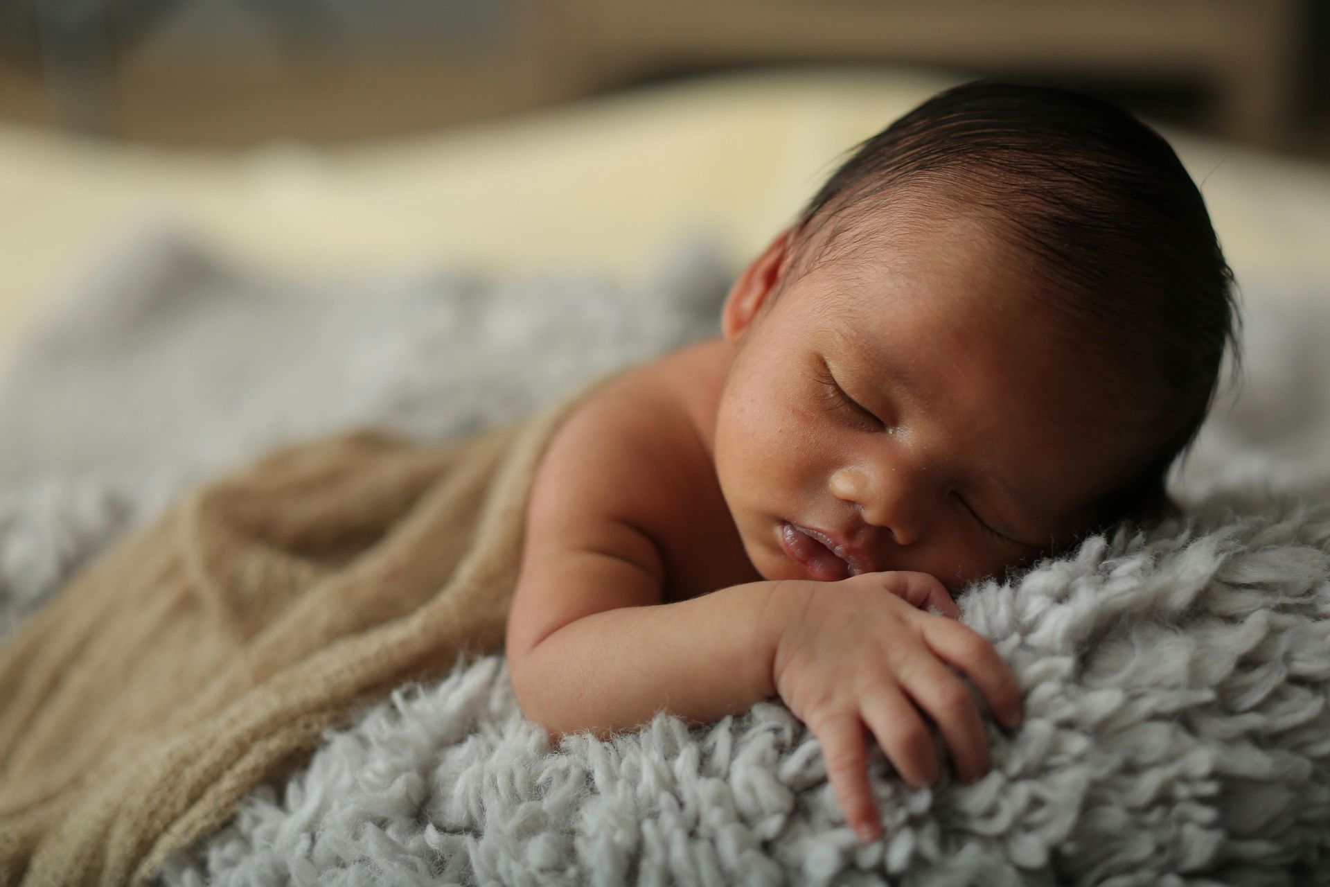 A newborn baby sleeping peacefully on a soft, grey textured blanket with a beige cloth draped over their shoulder.