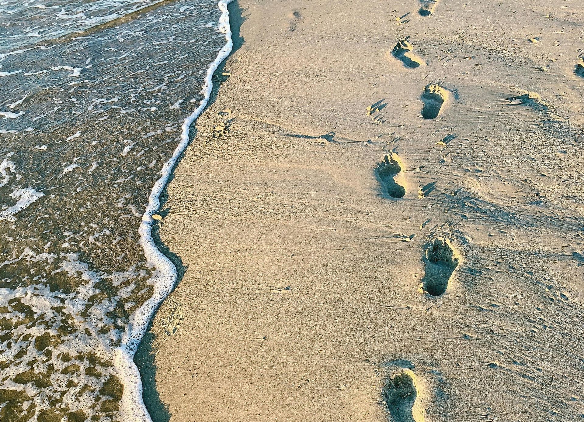 Footprints in sand next to foamy ocean waves.