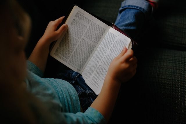 A child is sitting on a couch reading a bible.