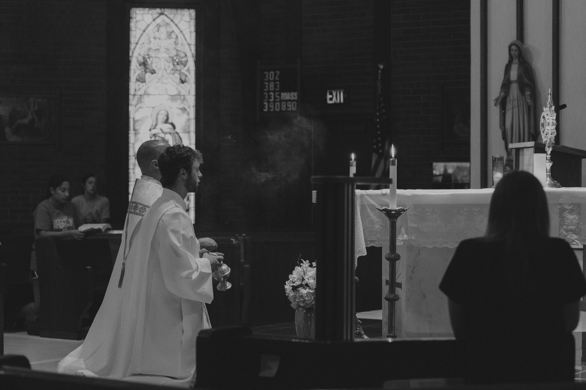 A priest and altar server kneel at a church altar. A woman prays nearby.
