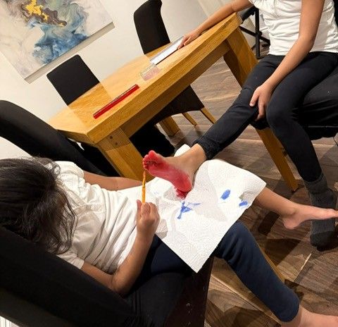 Two children painting one's foot red with a paintbrush, sitting at a table with art supplies.