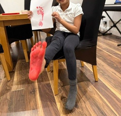 Child holding a red footprint art piece with a painted foot, seated on a chair.