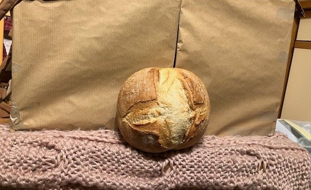 Round loaf of bread on pink knit blanket in front of two brown paper-wrapped items.