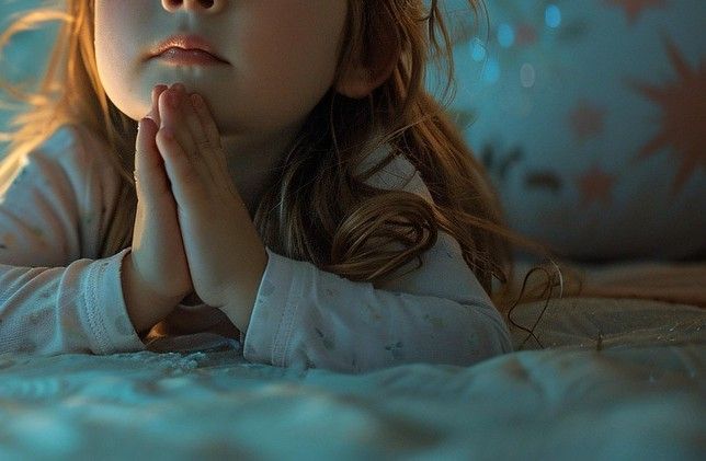 Child with hands clasped in prayer, resting on a bed. Soft lighting.