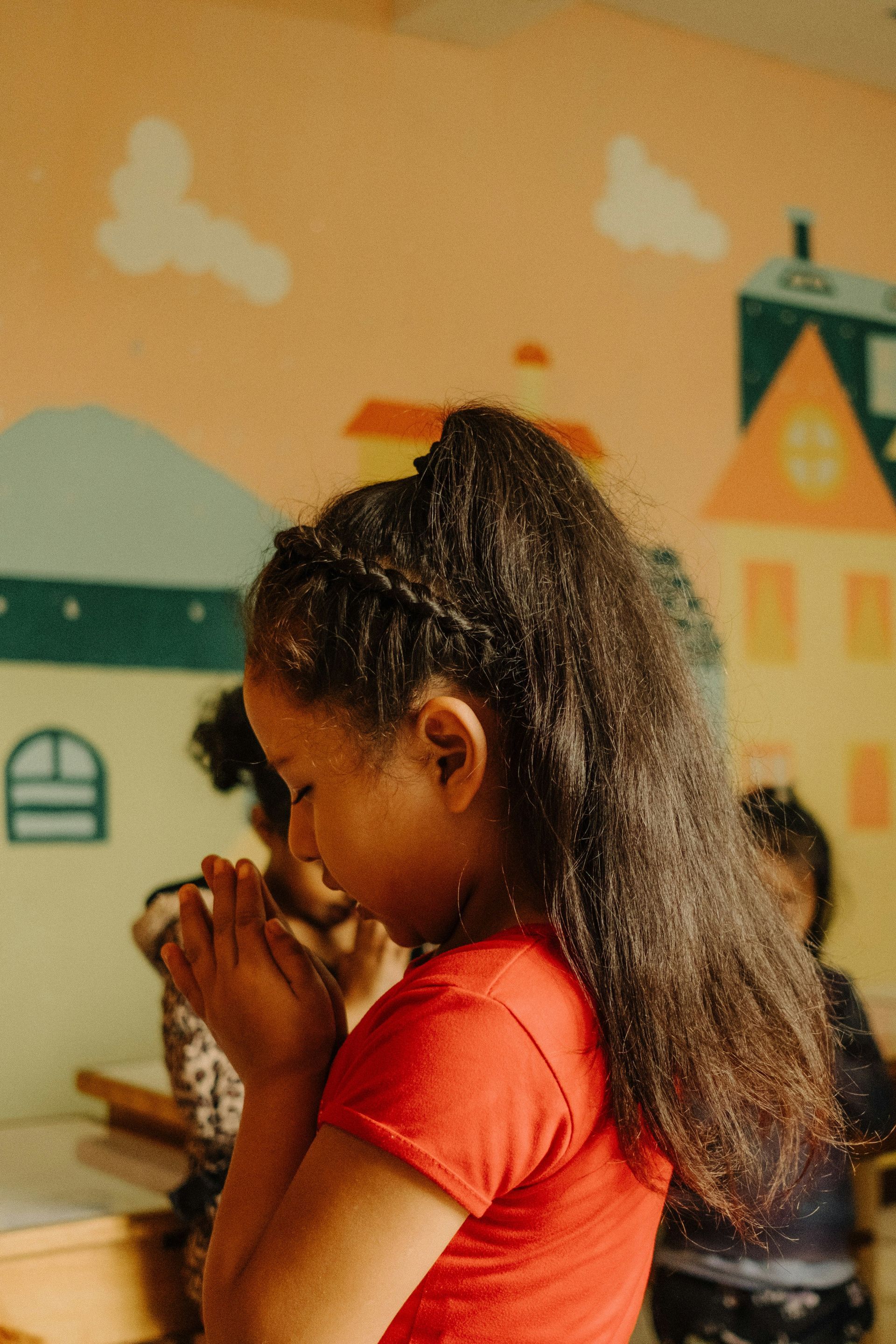 Girl with brown hair in a red shirt, praying with hands clasped, in a room with painted wall murals.