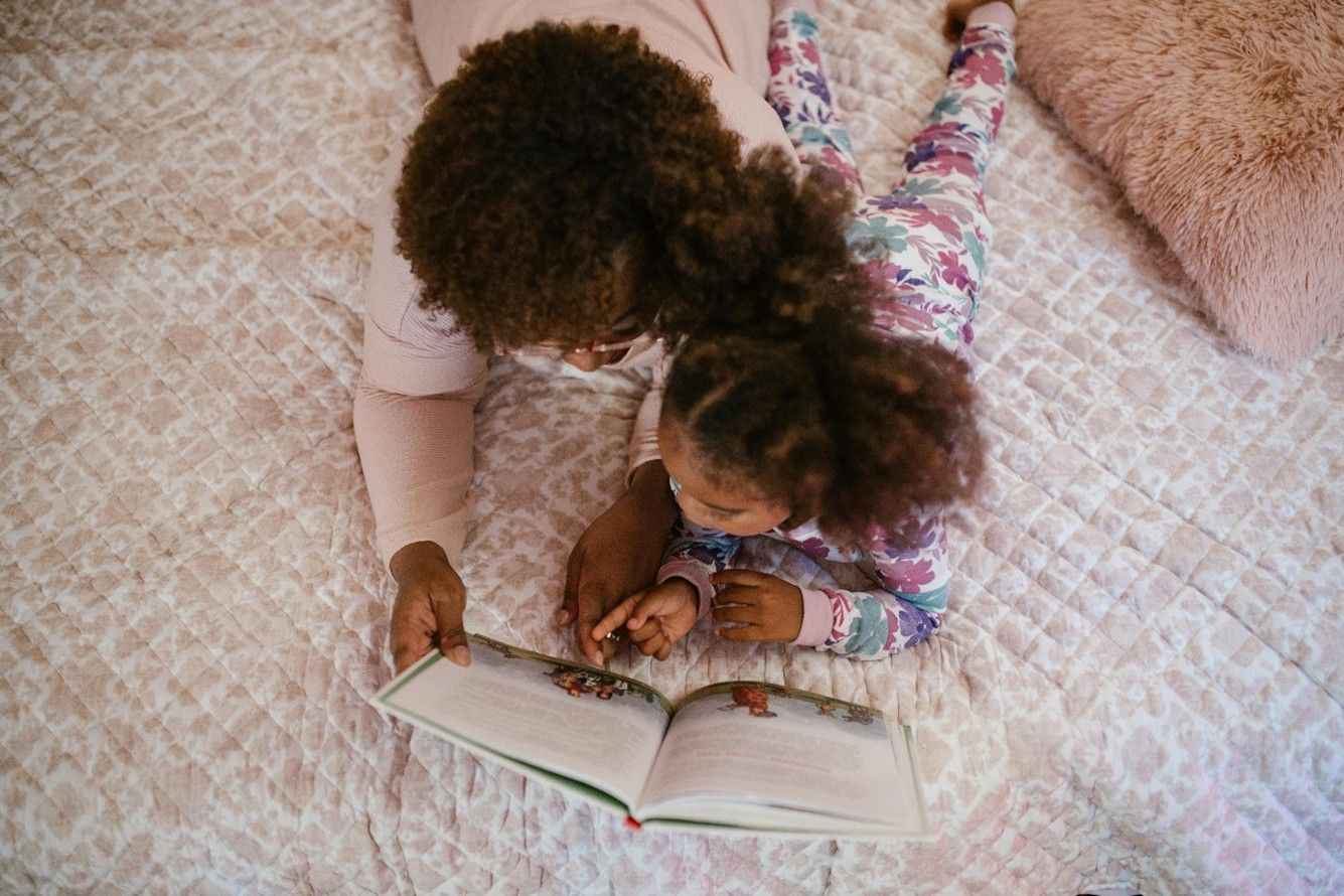 A mother and daughter are laying on a bed reading a book together.