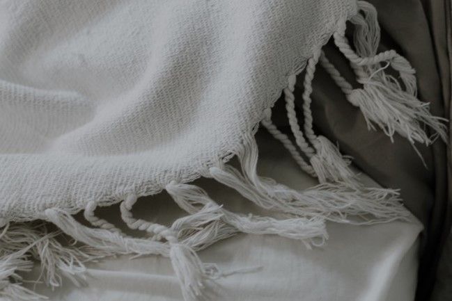 A white blanket with fringe is sitting on a bed.