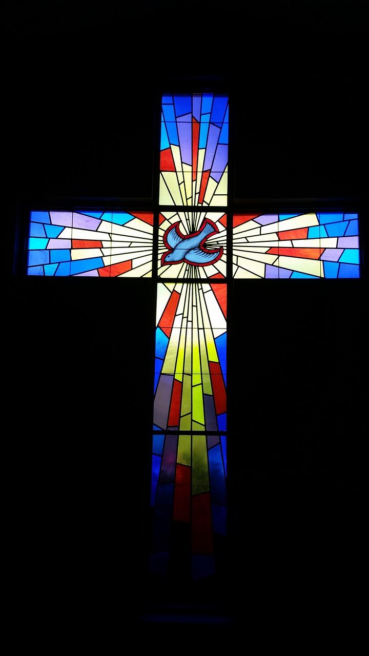 Stained glass cross with a dove in the centre, radiating colourful light against a black background.