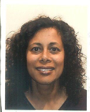 A headshot of a smiling person with shoulder-length, wavy, dark hair against a plain, light-colored background.