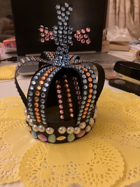 Black crown-shaped beaded decoration with pink, blue, and pearl accents on a yellow lace tablecloth