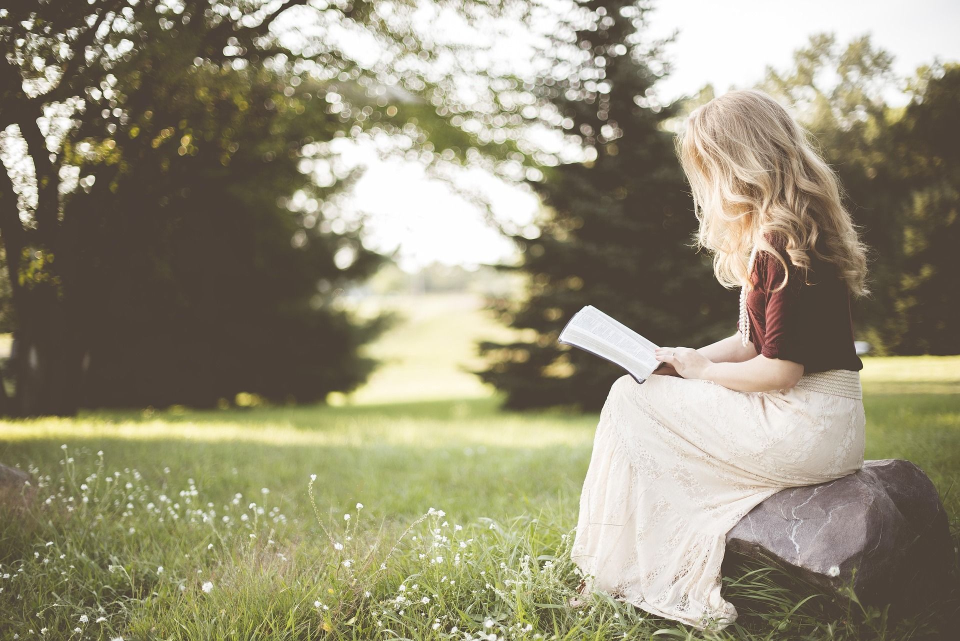 A woman is sitting on a rock in the grass reading a book.