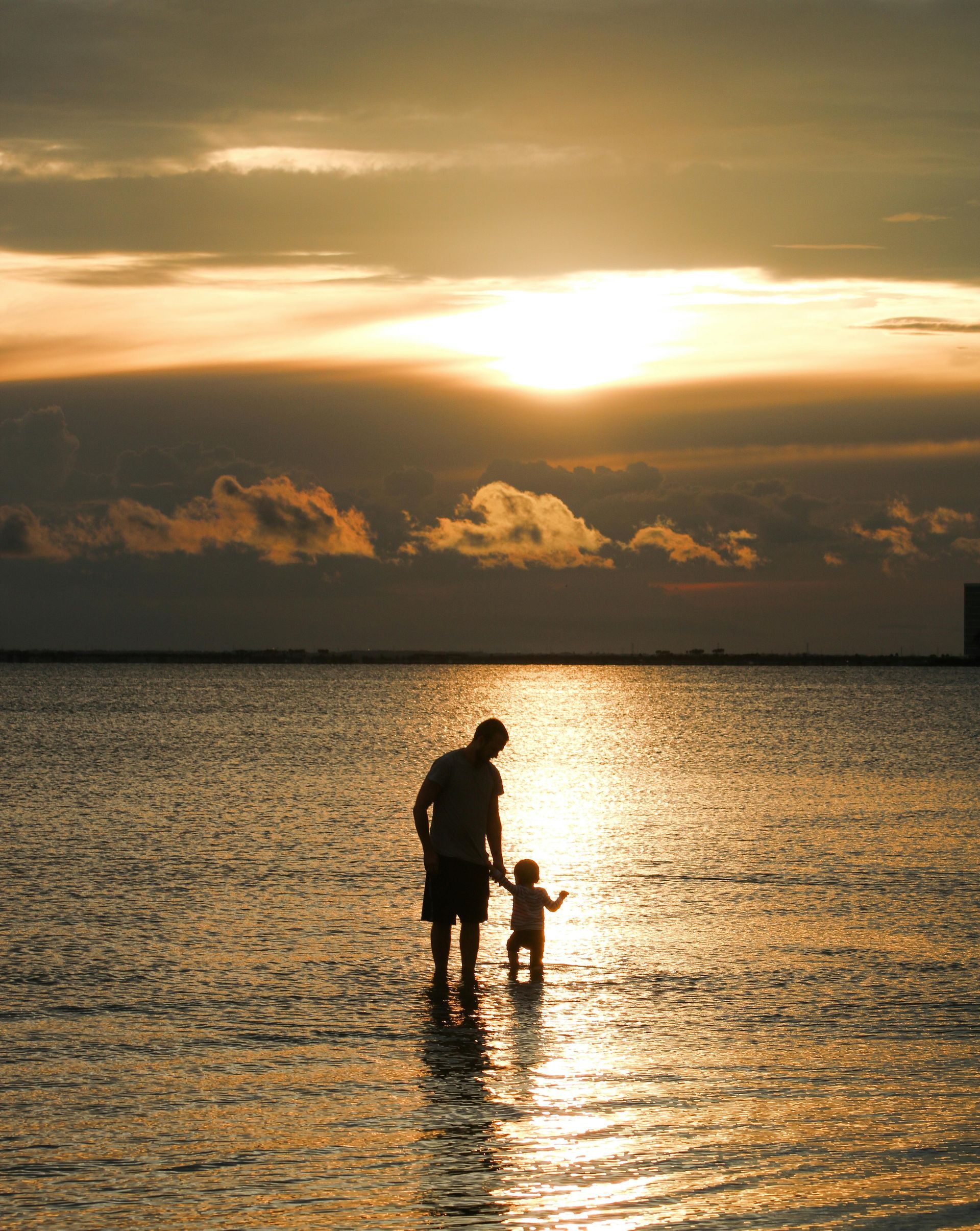 A man and a child are standing in the water at sunset.