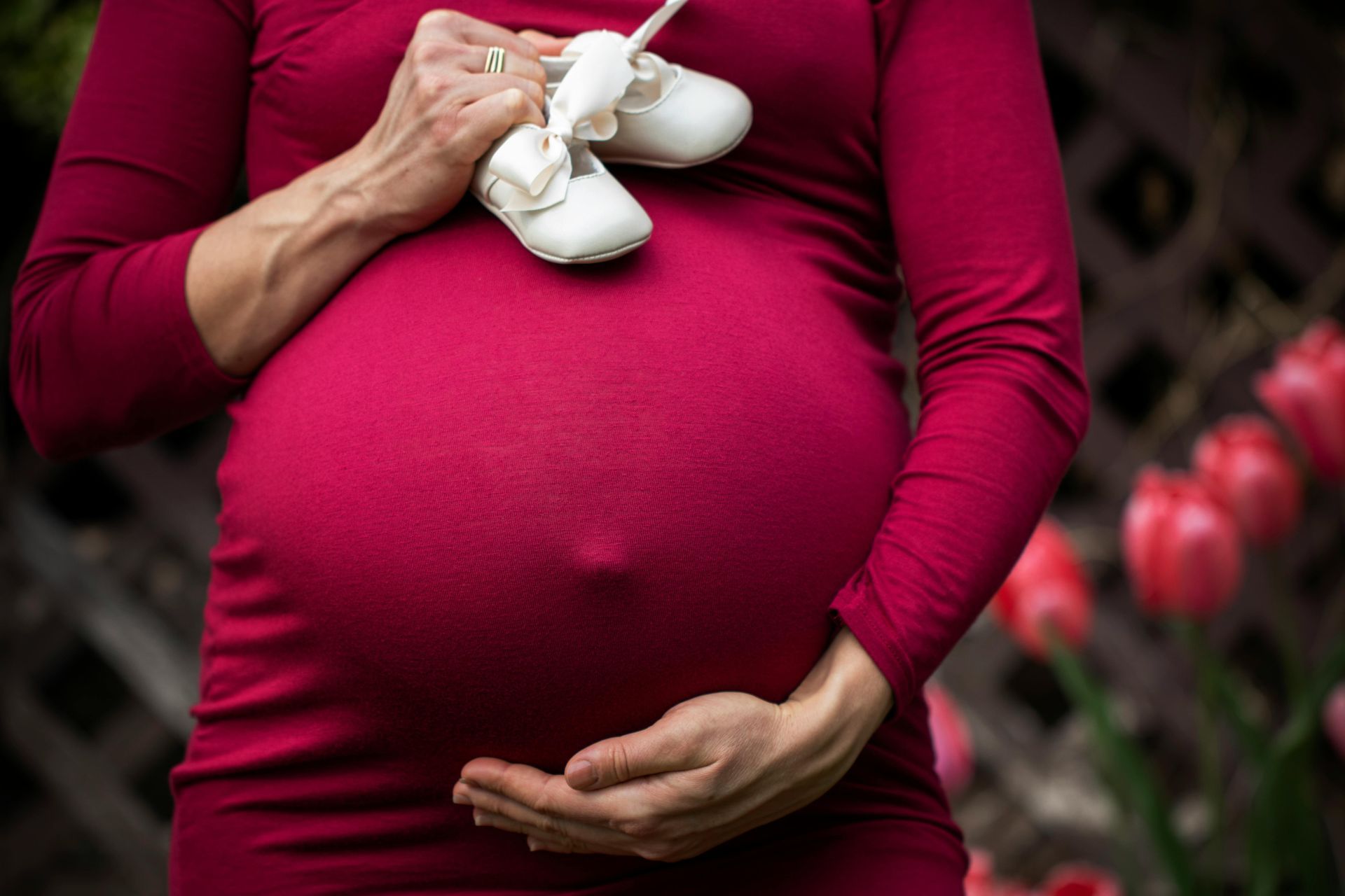 A pregnant woman in a red dress is holding a pair of baby shoes.