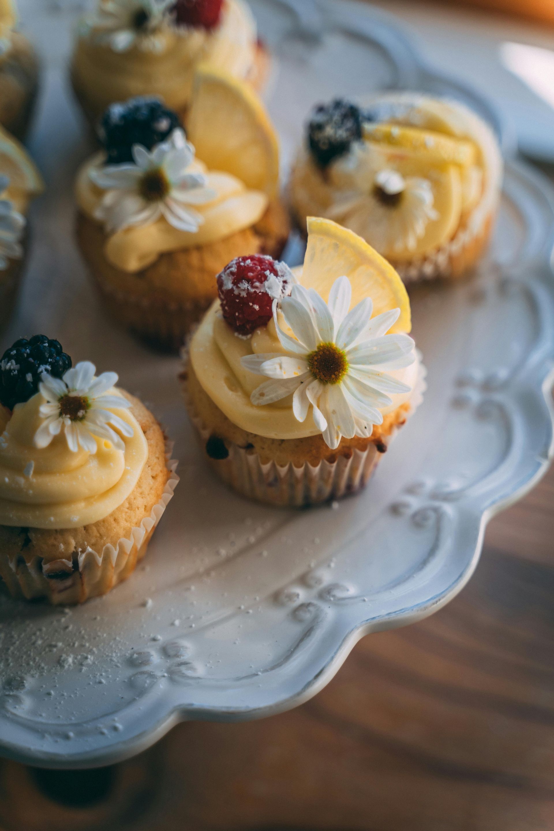 Cupcakes with yellow frosting, berries, lemon slices, and a daisy on a white decorative plate.