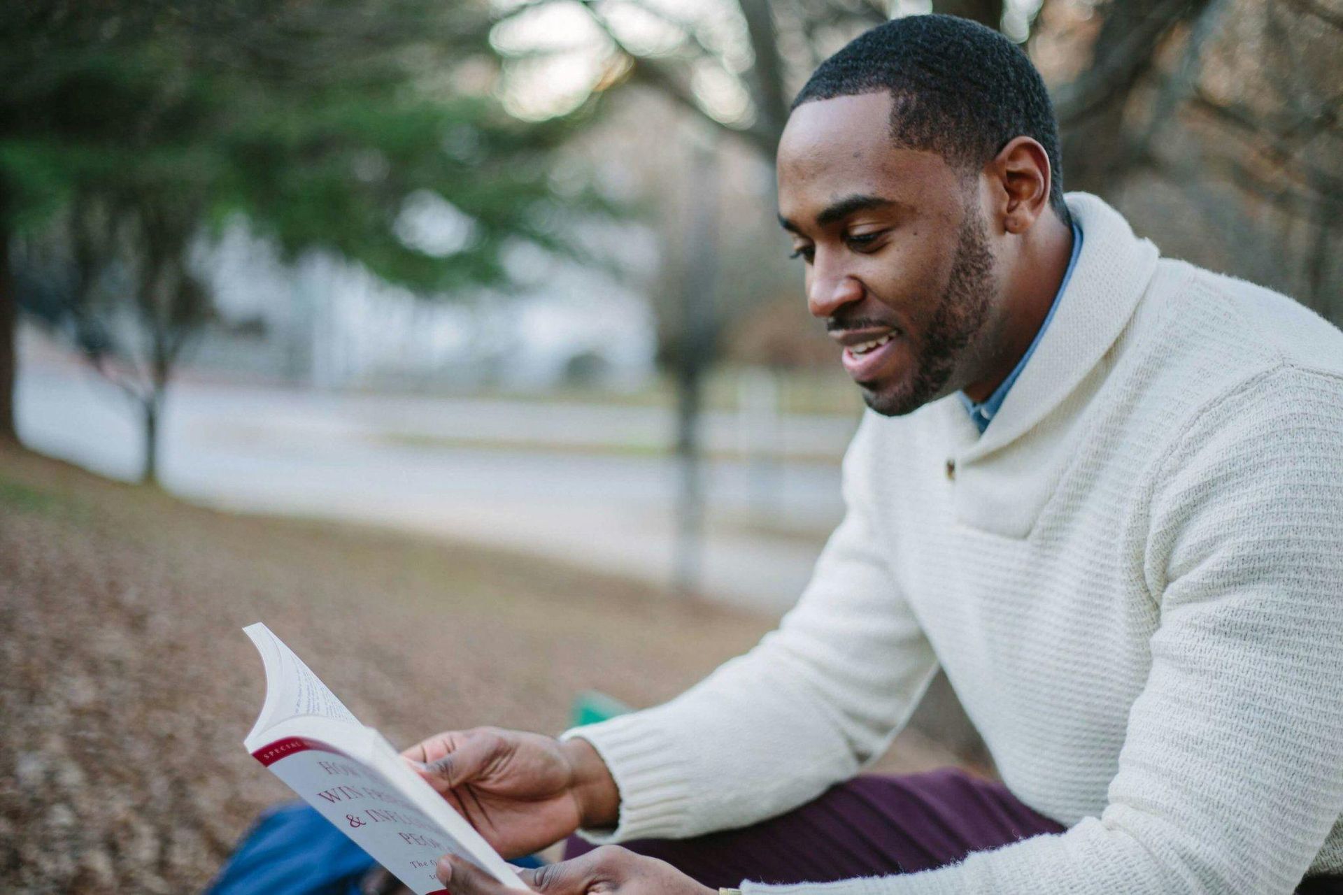 A man in a white sweater is sitting on the ground reading a book.