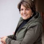 A woman wearing a green jacket and a black turtleneck is smiling.