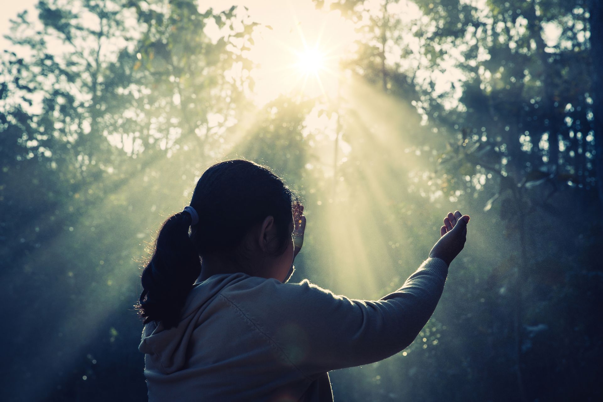 A woman facing sunlight, arms outstretched in a forest, rays of light shining.
