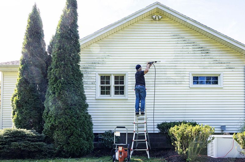 A man is standing on a ladder washing the side of a house.