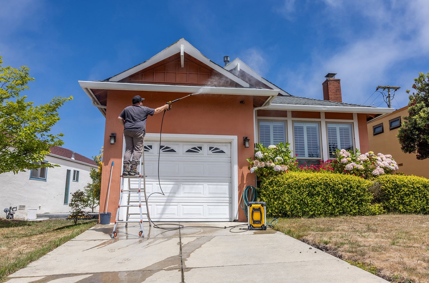 A man is standing on a ladder spraying a house with a high pressure washer.