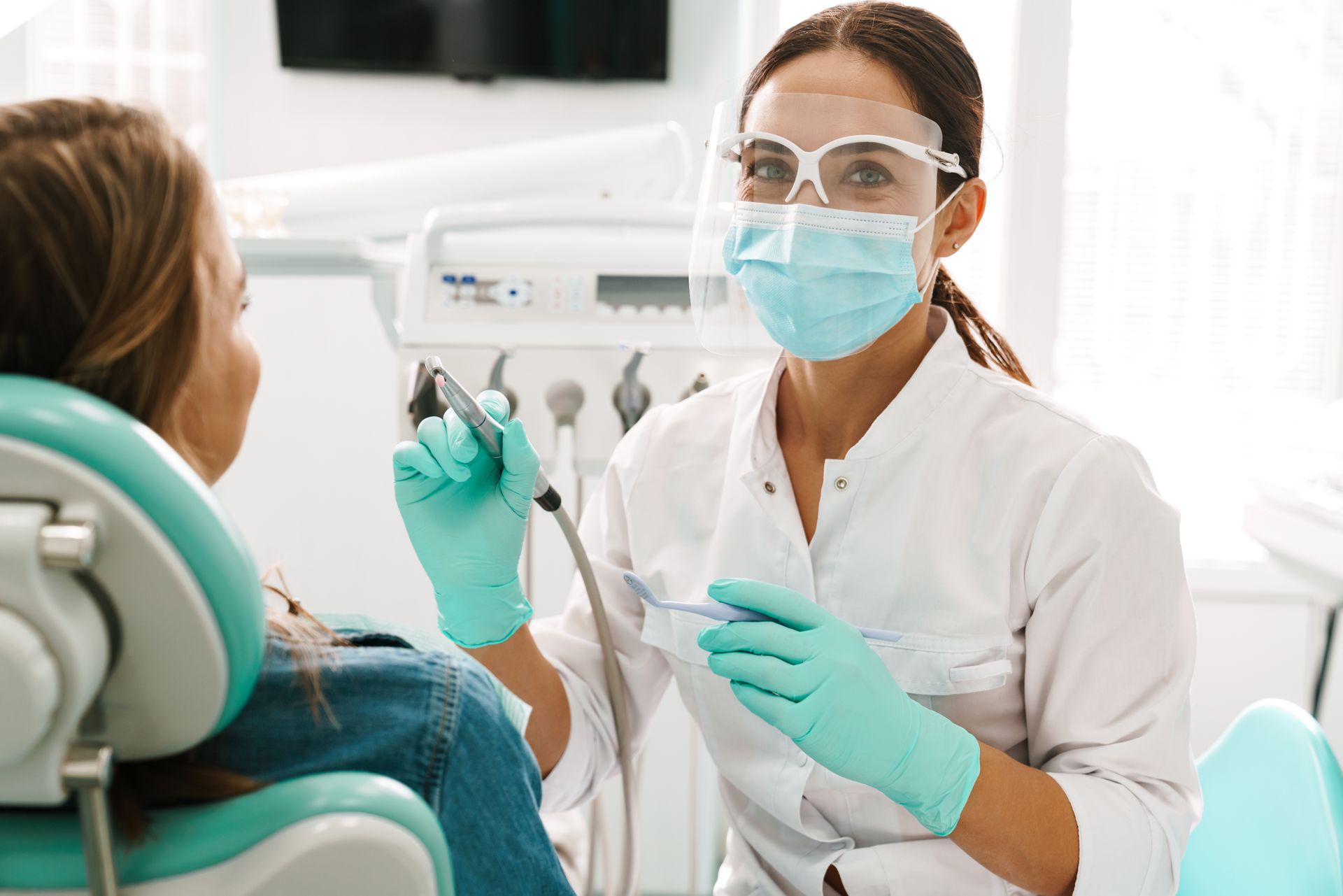 Dentist wearing gloves, mask, and face shield holding tools beside patient.