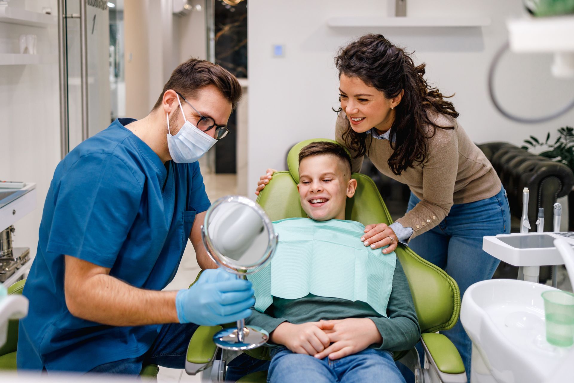 A family dentist holds a mirror for a boy to see his teeth while his mother watches contentedly.