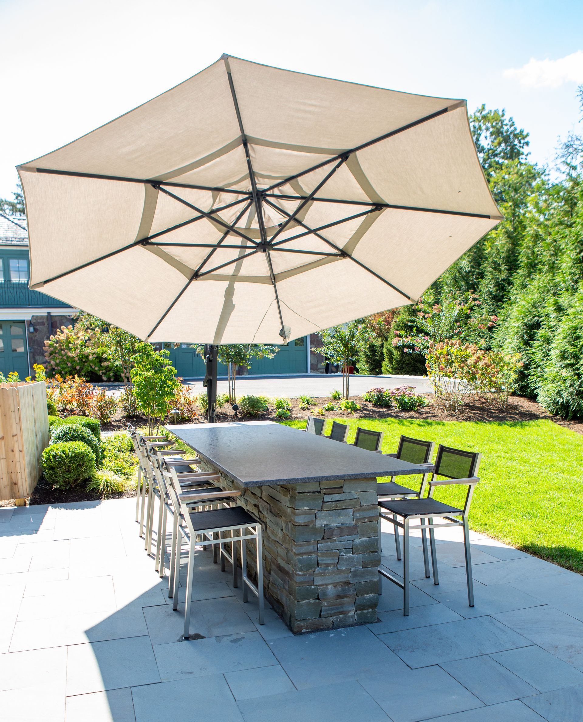 Patio dining set under large umbrella, stone table, metal chairs on paved surface.