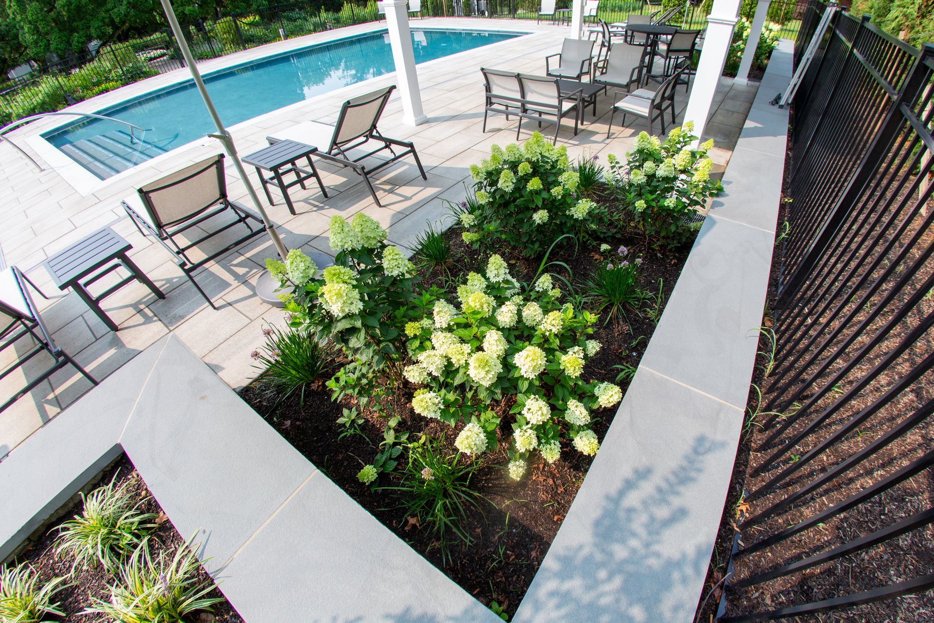 Poolside garden bed with white flowers next to a patio with seating and a pool.