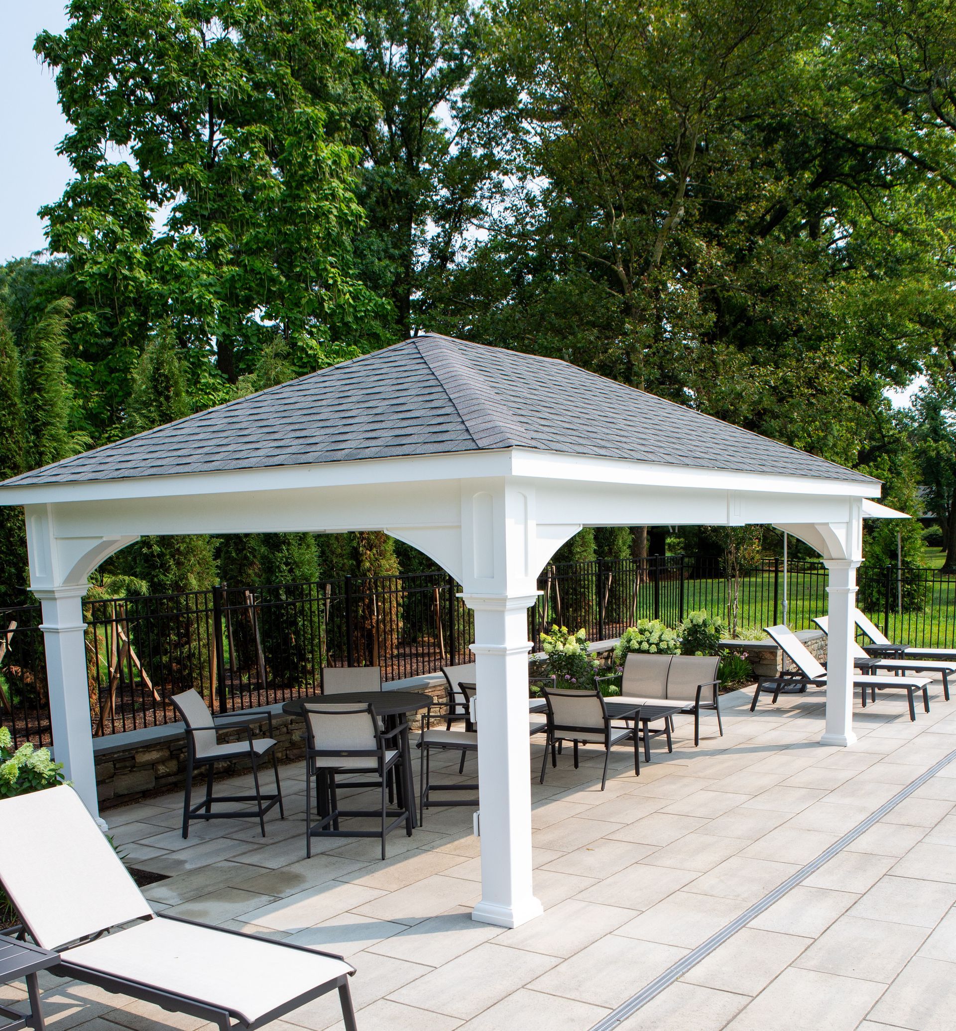 White gazebo with tables and chairs on a patio, surrounded by greenery, next to a pool.